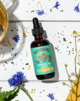 Bottle of Poppy Dream Elixir on white table with dried herbs and flowers
