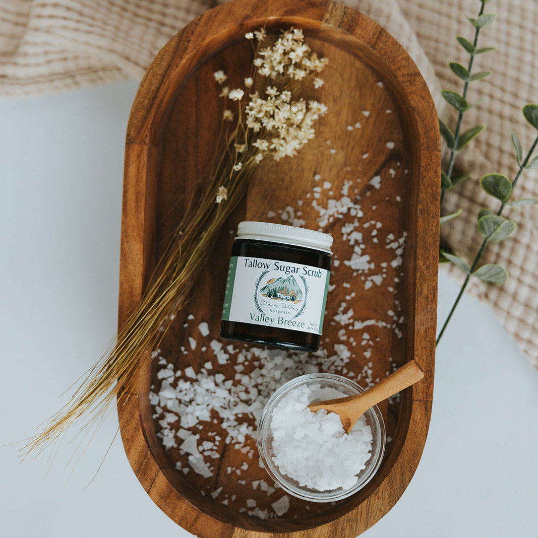 Wooden tray with a jar of sugar scrub, a small bowl of salt, and a wooden spoon on a light background.