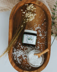 Wooden tray with a jar of sugar scrub, a small bowl of salt, and a wooden spoon on a light background.