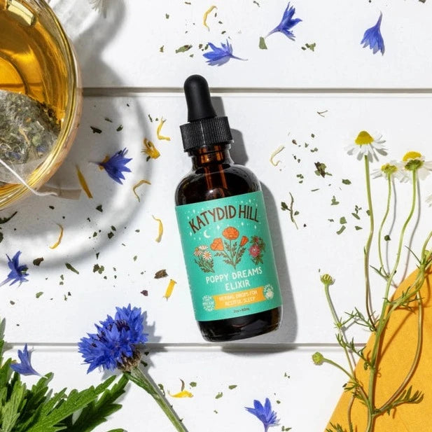 Bottle of Poppy Dream Elixir on white table with dried herbs and flowers