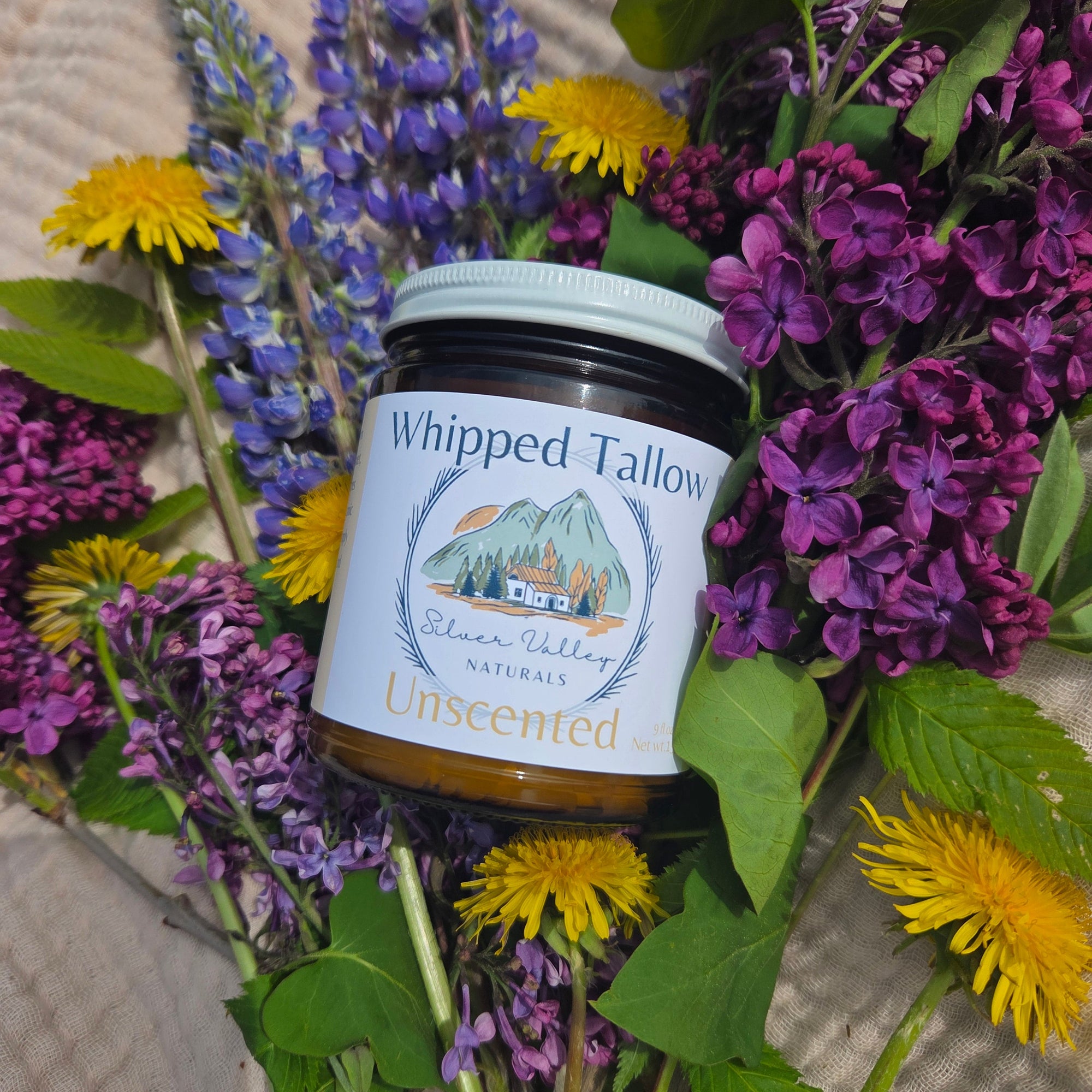 Jar of whipped tallow nestled among purple and yellow flowers on a textured surface